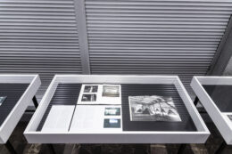 Zdjęcie przedstawia trzy gabloty, fotografowane z góry, ale tylko zawartość środkowej mieści się w kadrze w całości. Pozostałe są przedstawione jedynie w małych fragmentach. W środkowej gablocie leżą rozłożone wydruki i otwarte publikacje.