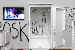 Zdjęcie przedstawia fragment ekspozycji w dużej, górnej sali galerii. W jego centrum znajduje się wysoka, pusta, otwarta lodówka bez półek. Na wewnętrznej ścianie został wykonany rysunek czarną, grubą linią, przedstawiający dwie ludzkie postaci: stojącą kobietę i klęczącego przed nią mężczyznę. Nad nimi świeci uśmiechnięte słoneczko. Rysunek przypomina dziecięce obrazki. Na lewo od lodówki wisi na ścianie ekran z projekcją.