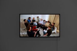 Fotografia przedstawia ekran z projekcją filmu Jany Shostak. Płaski ekran zawieszony jest na popielatej ścianie. Uchwycony na zdjęciu kadr z filmu przedstawia grupę 11 osób rozmawiających ze sobą. Wśród nich jest również artystka Jana Shostak. Osoby są w różnym wieku, kolorowo ubrane. Siedzą w kole, bardzo blisko siebie, na białych, niskich krzesełkach lub szarej kanapie.